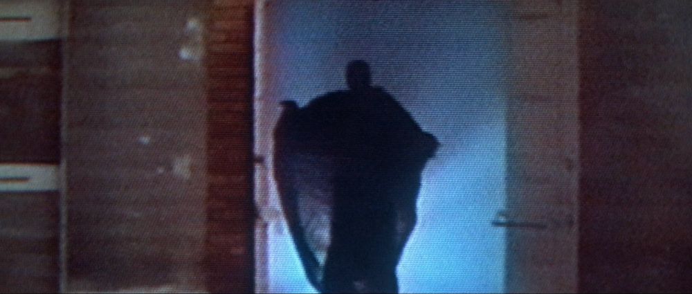 Prince of Darkness screenshot: grainy VHS quality image, a dark cloaked figure reaching out in silhouette against blue backlighting, emerging from a church.