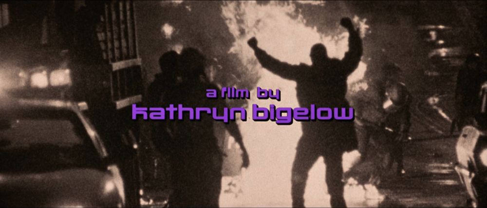 screenshot from Strange Days that reads 'a film by Kathryn Bigelow' in purple font over an image of a riot - a giant fire in the middle of the street, a police car at the left edge of the frame, the silhouette of a man with his fists raised and cops in the background. 