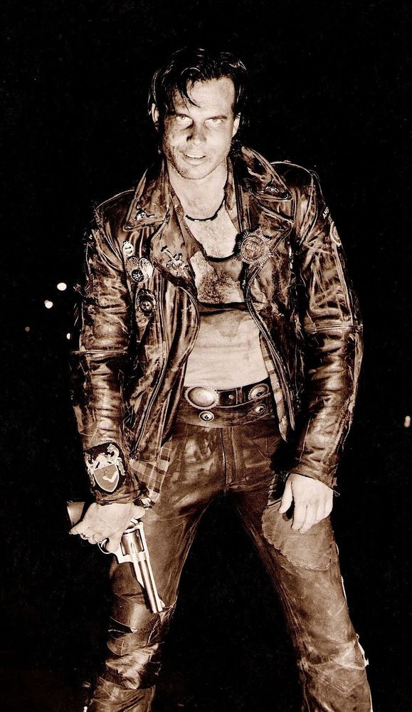 sepia-tone photo of Bill Paxton as Severen in Near Dark - looking bloodied and battered, holding a gun, wearing a distressed leather jacket with a sheriff star (& misc pins) and leather pants