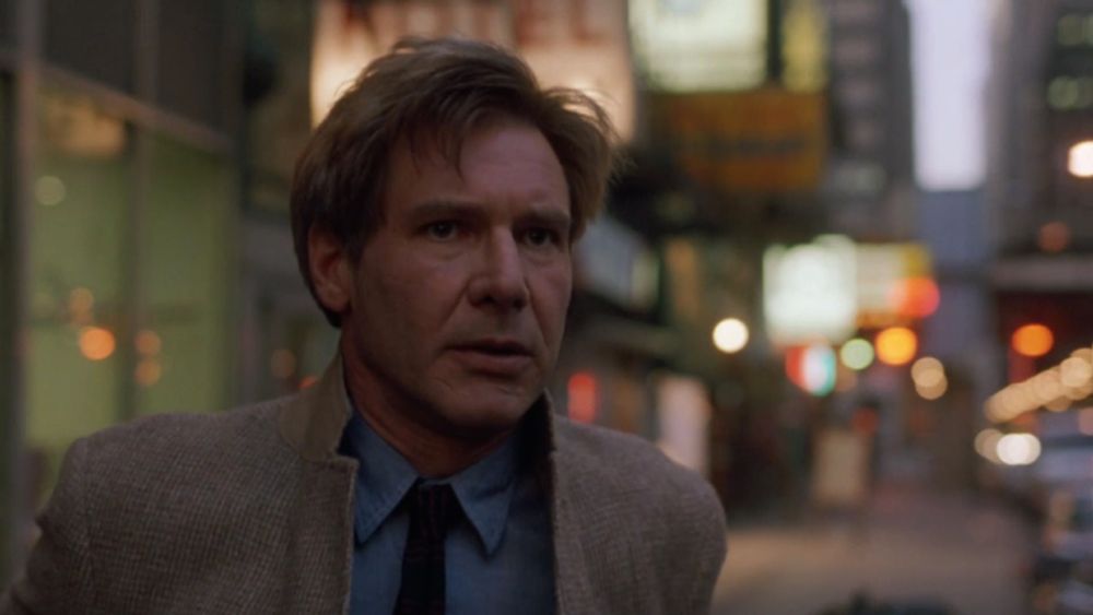 closeup on Harrison Ford in The Fugitive, standing on a sidewalk, the city behind him is out of focus. he's wearing an outfit that's giving Redford in Three Days of the Condor - tweed jacket with popped collar, a chambray shirt, and a black skinny knit tie (with tiny red stripes that are impossible to make out here) 