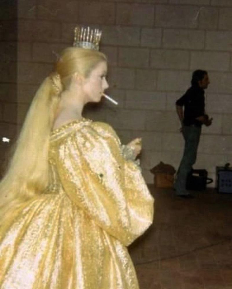 slightly fuzzy photo of Catherine Deneuve standing up, in profile, wearing an enormous gold, textured gown with poofy sleeves. her hair is pulled back, she has a long blond ponytail tied back with a braid of hair. there is a tiny crown on her head, a cigarette dangles from her mouth. a man on set stands in the background wearing contemporary cloths (long-sleeve shirt, jeans) 