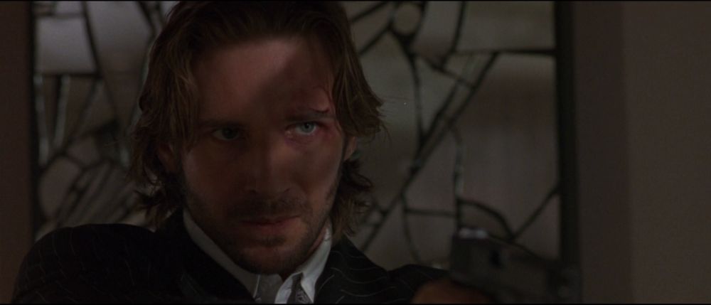 closeup on Fiennes against a broken mirror mosaic backdrop, he's a little bloody, wearing a pinstripe jacket, white shirt, paisley gray tie, and holding a gun. mostly in shadow but with a band of light over his eye (on our right) 