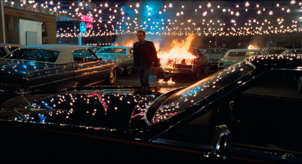 James Caan in the middle of a used car lot that's on fire. there's a pink and blue neon sign in the background and strings of white lights above that are reflected on the cars' surfaces, especially one black car that takes up most of the foreground 