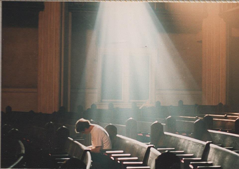 grainy photograph - a wide shot and side view of Wilson with shaggy hair in a white t-shirt sitting in church pews. a giant white light from the ceiling above surrounds him 