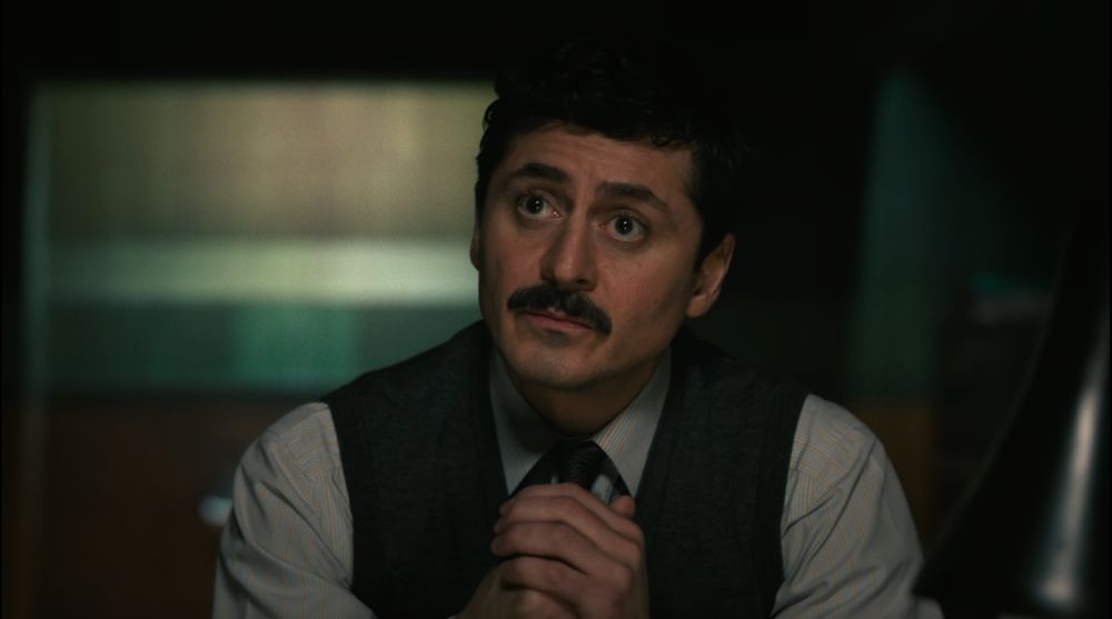 screenshot of Akram in Dept Q's pilot episode - handsome brunette, dark-eyed man with mustache, hands clasped in front of him, wearing a pale blue button-up shirt with tie and vest. against a fuzzed-out green backdrop 