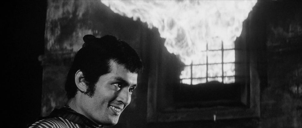 young evilly grinning Tatsuya Nakadai in Yojimbo, to the left of a window belching flames into the top of the frame 