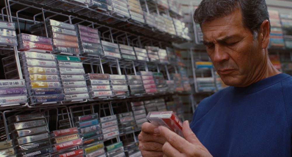 Max Cherry standing in front of shelves of cassette tapes, the shelves fill the frame. he's wearing a cobalt blue sweatshirt, his brow is furrowed as he studies the Delfonics tape in his hands 