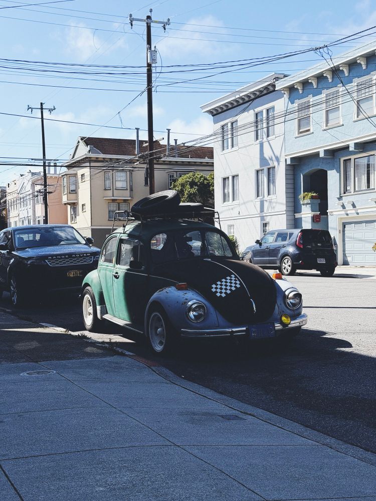 A classic Volkswagen Beetle with a two-tone black and blue color scheme and a small rectangular checkerboard decal on at an angle on the hood, parked on a San Francisco street. 