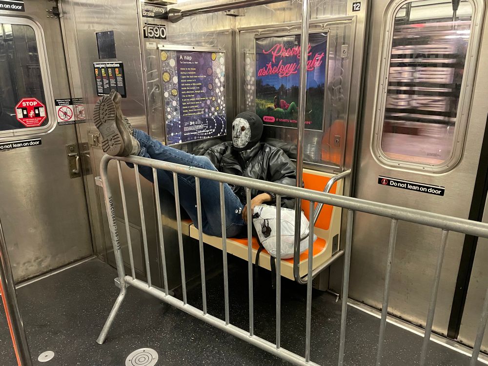 A masked man sits on the bench of an NYC subway car, with his feet up on a metal police barricade