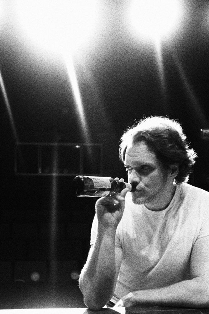 Stylish black and white photo of me leaning against a bar top and drinking from a prop beer bottle, looking menacingly off to the side. Stage lights dominate the top quarter of the photo.