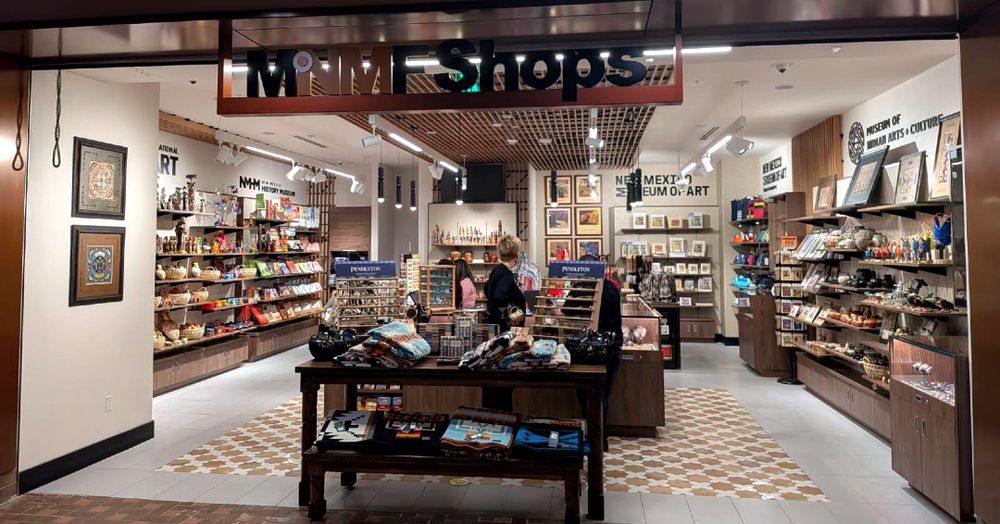 A souvenir shop with a woman browsing. Shelves display pottery, books, and art. A sign reads "New Mexico Museum of Art," highlighting local crafts.