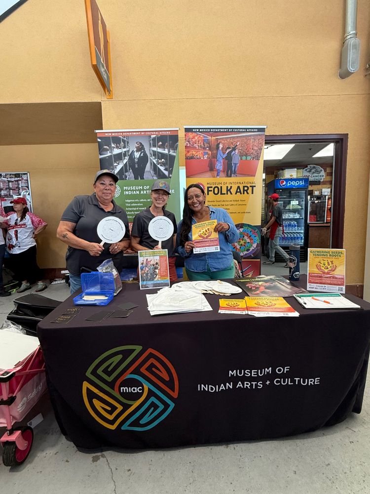 Three individuals stand behind a Museum of Indian Arts & Culture table, holding promotional materials at an event.