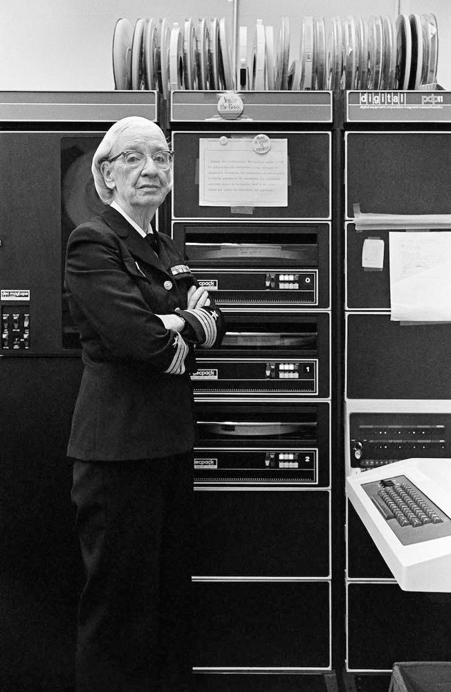 Rear Admiral Grace Murray Hopper, computer scientist, shown in black & white, in her navy uniform in front of an early computer cabinet. Her arms are crossed and she’s looking a bit sassy. 