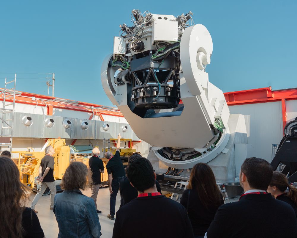 A crowd of onlookers visit ESA's new Flyeye telescope inside the factory where it is undergoing final testing. The roof of the factory has been rolled back to offer the telescope a clear view of the sky.