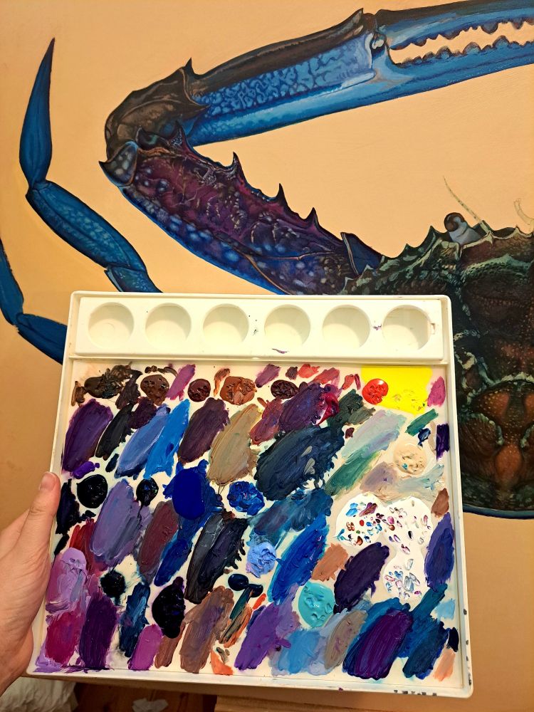 A photo of a crab painting on panel. There is a paint palette showing many colours the artist is using. The colours include bright blues, purples and pinks.