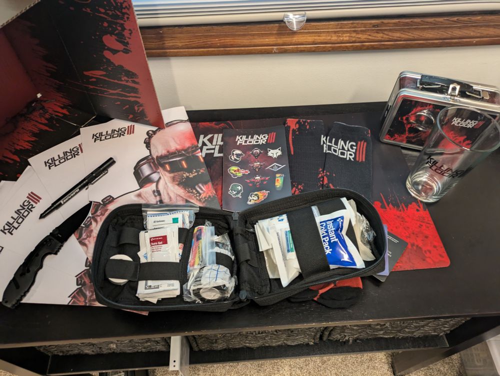 A variety of Killing Floor 3-branded items, including a lunch box, a pint glass, a first aid kit, dress socks, stickers, a mouse pad, a tactical knife, an emergency pen, and shooting targets