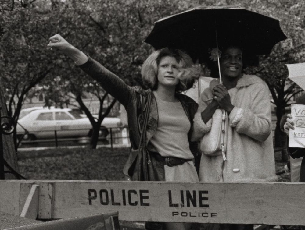 Sylvia Rivera and Marsha P. Johnson stand beneath an umbrella, behind a barricade stenciled "Police Line".