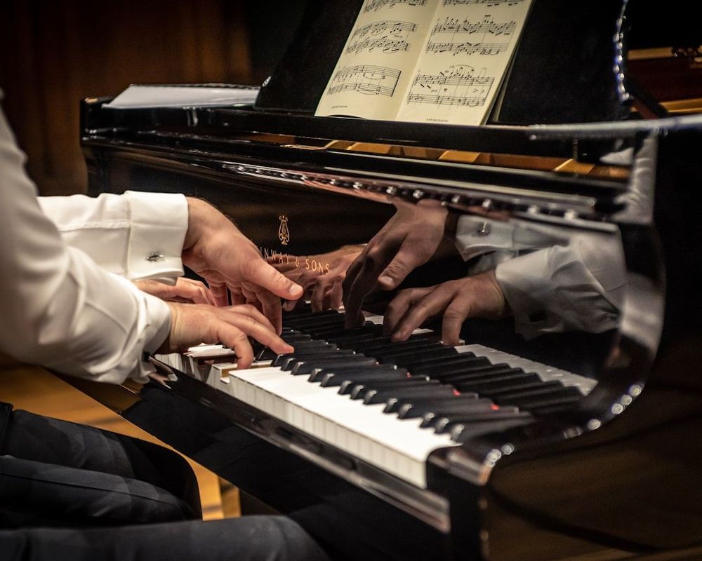 Photo of someone playing a black grand piano. Only the player's hands are visible. There appears to be someone seated next to them. They are reading from sheet music and the player's hands appear to be in a graceful pose. 