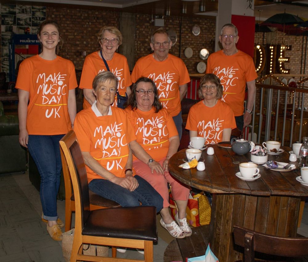 A group of seven people sits and stands around a wooden table in a café, wearing matching bright orange "Make Music Day UK" T-shirts. They smile at the camera, with cups of tea and plates on the table. The café features warm lighting and eclectic decorations, including wall-mounted plates and a vintage police box in the background.