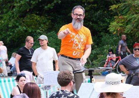 A conductor wearing an orange "Make Music Day UK" t-shirt leads a group of musicians during an outdoor performance in a park. Several people watch or pass by in the background, with trees and striped deckchairs visible.