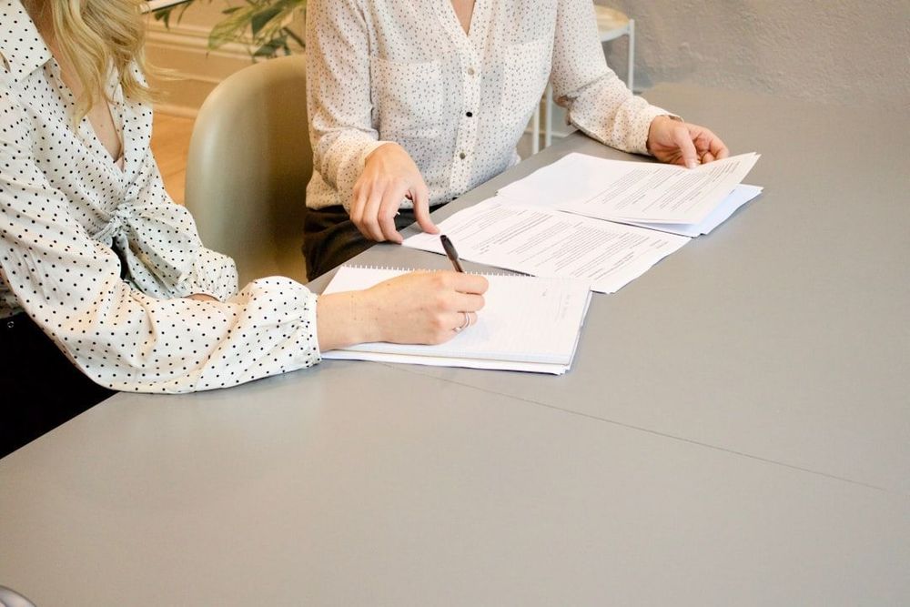 Two people sitting at a table reviewing documents and writing notes, with printed papers and a notebook spread out in front of them. Both are wearing polka dot blouses.