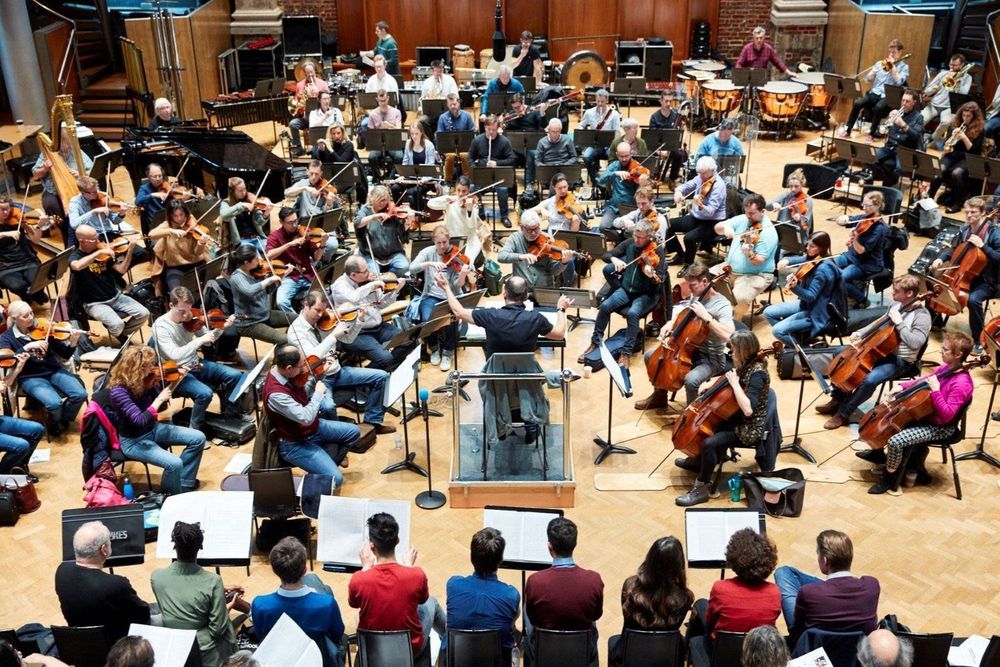 A large orchestra in rehearsal, viewed from above and behind the conductor. The conductor stands on a podium with arms raised, leading a full ensemble of string, woodwind, brass, and percussion players. Several people sit in the foreground with open scores, observing.