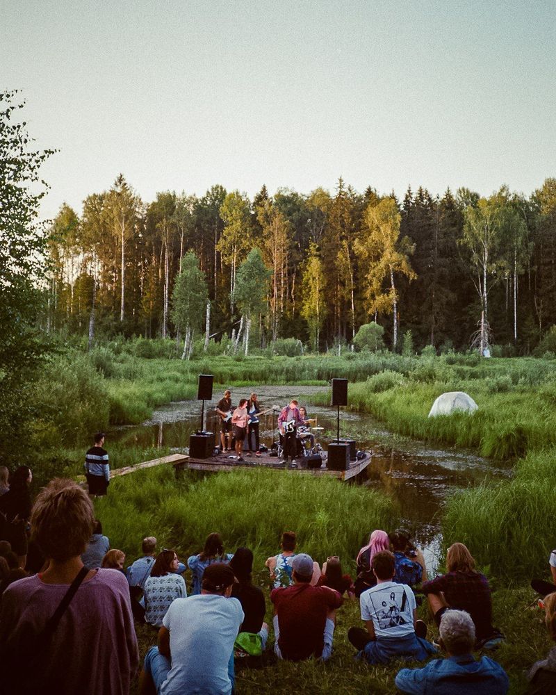 A photo of a group of people sitting in nature listening to a music group performing. There is a forest in the background and it appears to be sunset. It has a peaceful and tranquil feel to it. 
