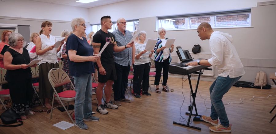 A choir rehearsal in a community hall, where a diverse group of older and middle-aged singers stands holding sheet music, following along with the conductor. The conductor, a man wearing a white shirt and jeans, leans forward while playing an electric keyboard, passionately leading the group. The singers appear engaged, focused, and expressive. The room has wooden flooring, chairs with red cushions, and natural light coming in through small windows.