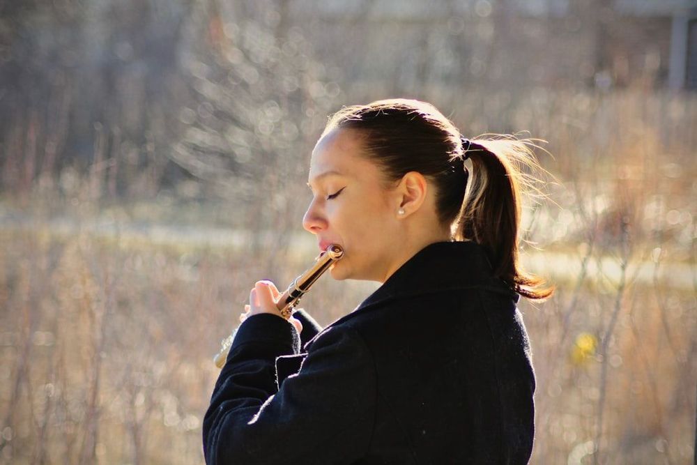 A women playing a flute in nature. She appears to be deeply engrossed in her playing. 