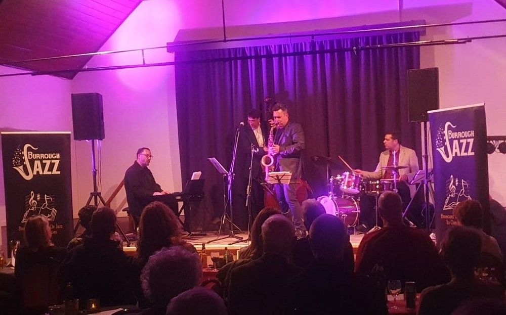 A jazz band plays on stage in a small dimly lit venue. The audience can be seen in the foreground. There is a keyboardist, saxophonist, double bassist, and drummer on stage. There are signs on either side of the stage with the band's details on.