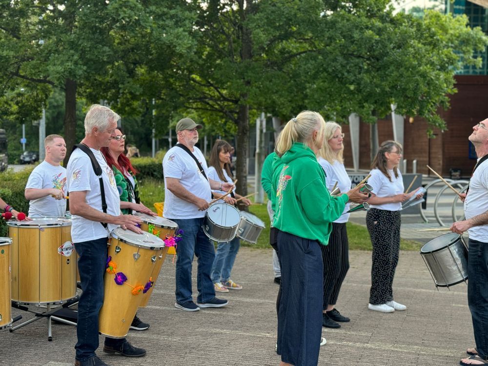 A group of people dressed in white t-shirts and casual clothes play various percussion instruments, including drums and tambourines, in an outdoor urban setting with trees and modern buildings in the background. Some participants wear green hoodies with a logo on the back.