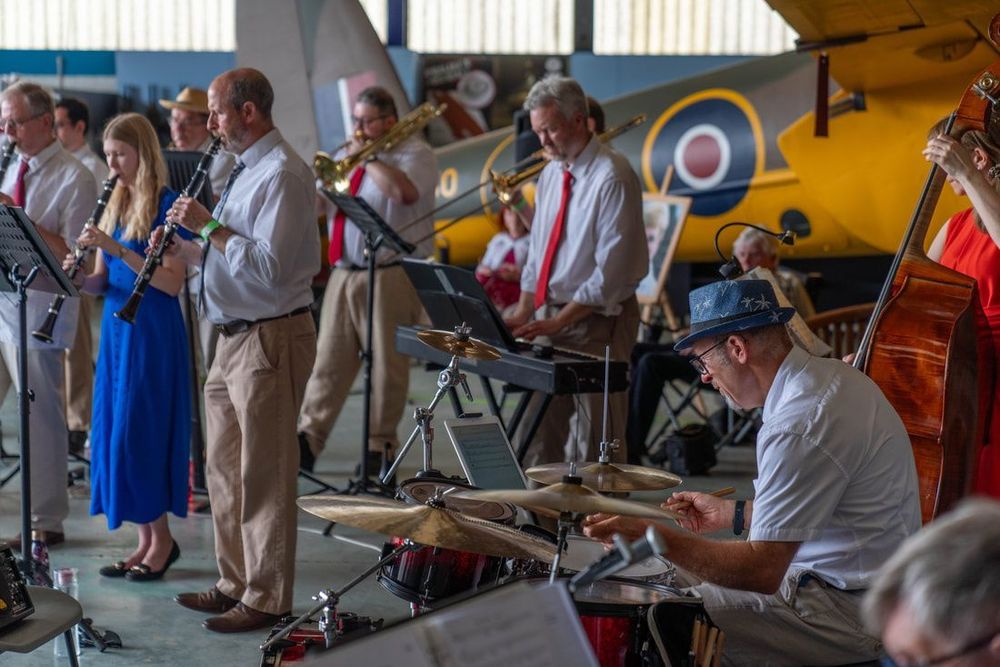 A swing band performs in front of a vintage aircraft in a hangar. Musicians play clarinets, trombone, keyboard, double bass, and drums. They wear smart casual outfits, with many in shirts and ties. A woman in a bright blue dress plays clarinet. The setting has a lively, retro feel.