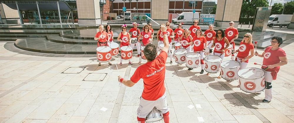 A group of people in matching orange shirts and white trousers play drums in a semi-circle outdoors, led by a conductor with drumsticks raised. They are performing in a public square near modern buildings and a water feature, under bright sunlight.