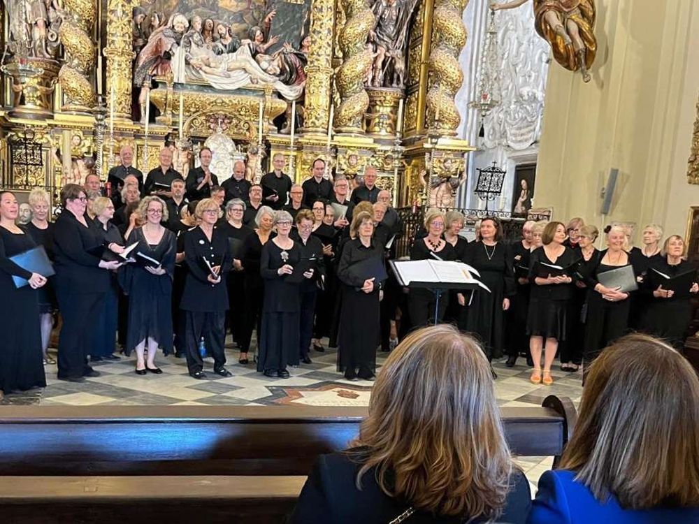 A large choir dressed in black performs in an ornate church, standing in front of an elaborate gilded altarpiece with religious artwork and sculptures. Two audience members are visible in the foreground, seated in pews, watching the performance.