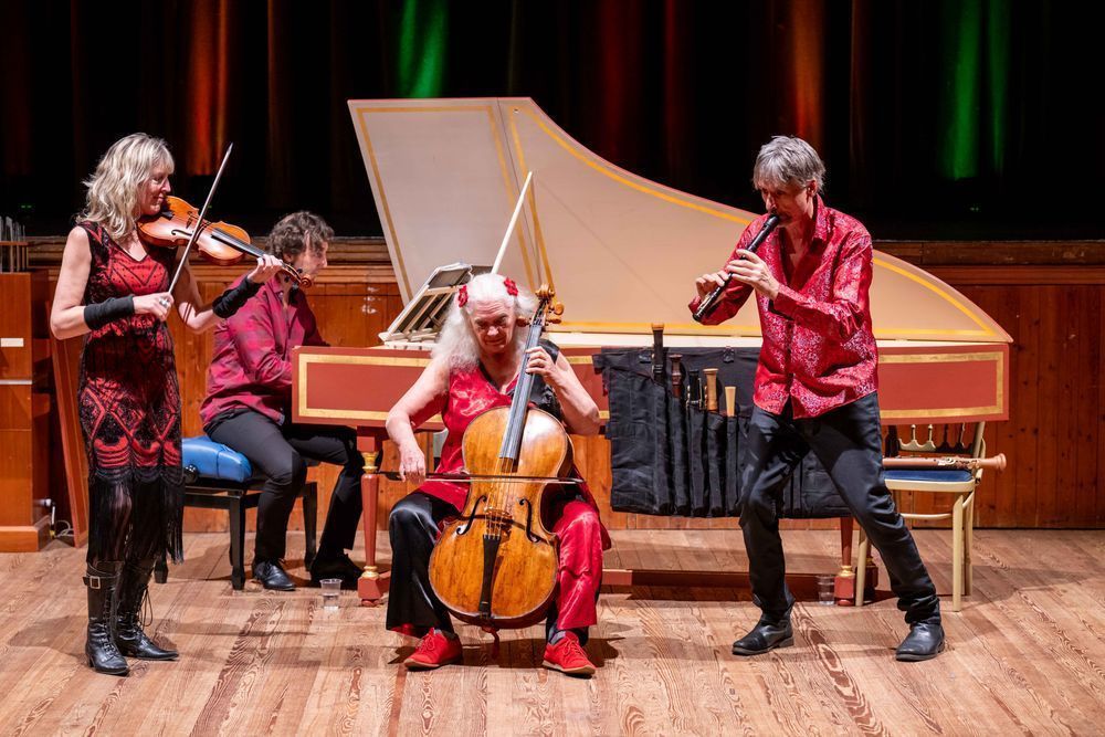 Four musicians perform on a wooden stage in vibrant red and black outfits. A woman plays the violin, a man plays the harpsichord, another woman plays the cello, and a man plays a recorder. Behind them is a grand harpsichord and a curtain lit with warm, colourful stage lights.