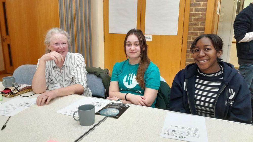 Three women sit at a table in a well-lit room, smiling at the camera. Papers, pens, and mugs are scattered on the table. One woman wears a green t-shirt with a music-themed logo, and another wears a dark blue hoodie with a similar logo.