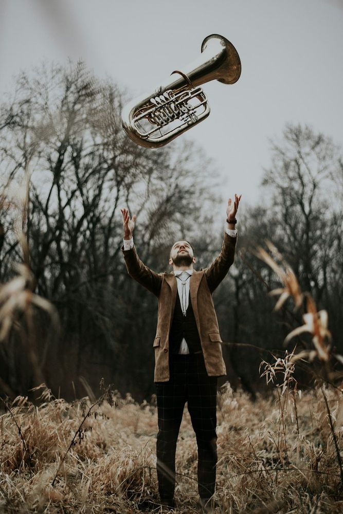 A photo of a man standing in nature. He is throwing a euphonium in the air. The atmosphere is joyful.