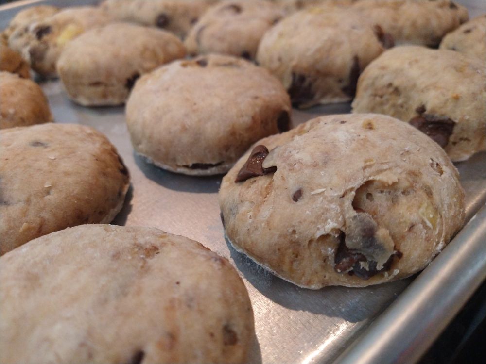 A pan of pleasantly puffy banana bread cookies with dark chocolate chips. The closest cookie, clearest in zoom, shows the flaky sides of well floured dough on the outside; however, the insides visible through the cracks look moist and tender. This is all, of course, the opinion of the woman who baked the cookies and wrote the alt text.