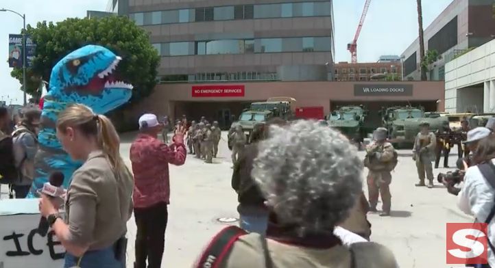 LA Roybal federal building. A bunch of journalists take photos of the mobilized national guard, who are just standing around like clowns. A person in a large blue dinosaur costume holds an anti-ICE protest sign.