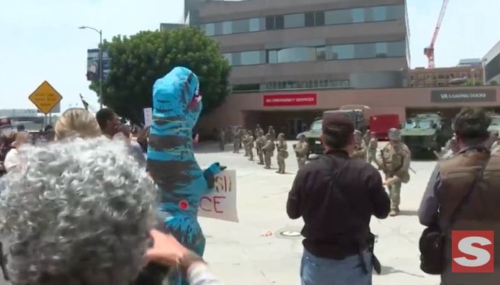 LA Roybal federal building. A bunch of journalists take photos of the mobilized national guard, who are just standing around like clowns. A person in a large blue dinosaur costume holds an anti-ICE protest sign.