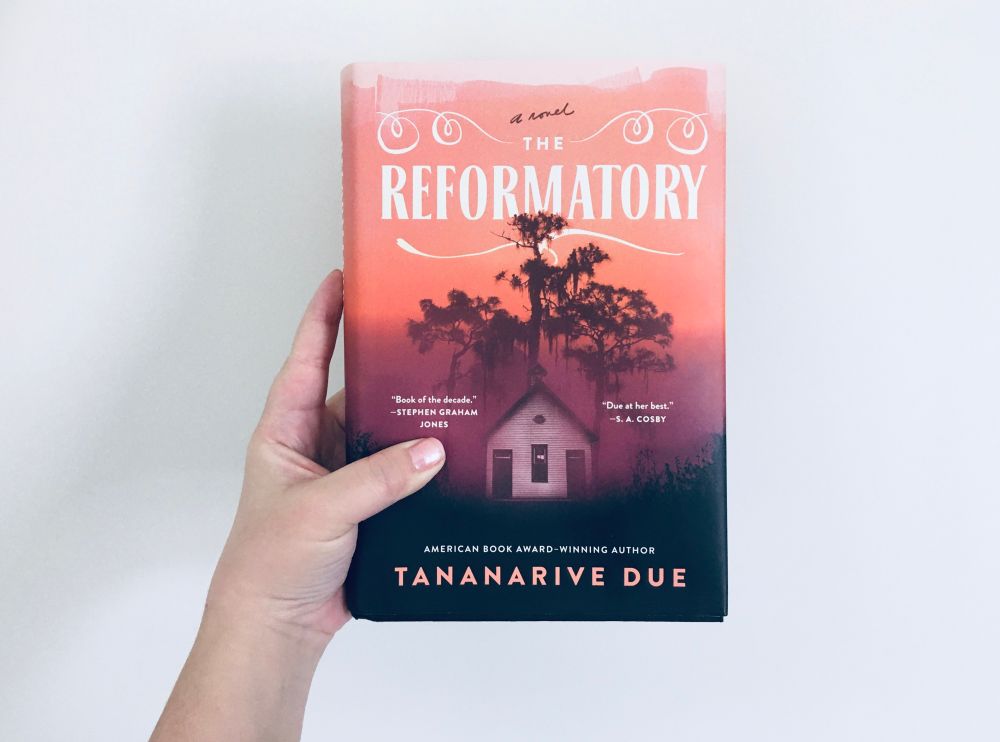 Me holding a copy of The Reformatory by Tananarive Due:
The cover shows an ominous shack.