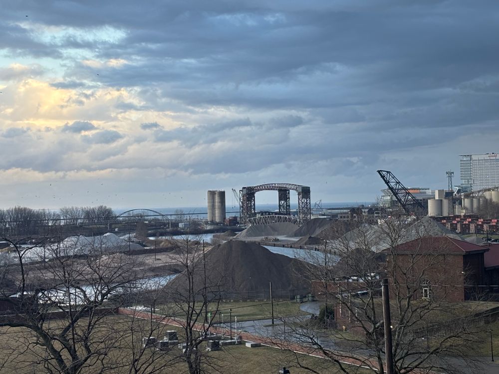 This is a photo of an industrial part of the near west side of Cleveland with some bridges, trucks and Lake Erie in the background. The sky is cloudy. 