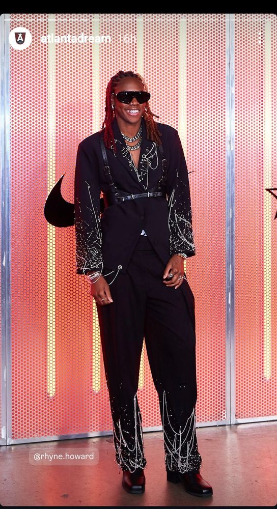 Rhyne Howard wearing a black suit with silver chains and embellishments along the lower legs of pants and towards the mid to end of suit jacket sleeves. Also wearing a black leather harness. Silver chairs and embellishments also on the lapels. Silver chain and rings bracelets. Black sunglasses