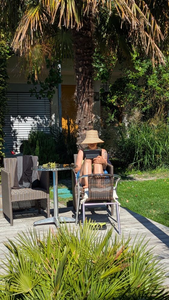 Eine Person sitzt lesend mit großem Sonnenhut auf einem von zwei Korbsesseln mit den Füßen auf einem Korbstuhl auf einem Holzpodest in einem Garten unter einer großen Palme. Zwischen den Sesseln steht ein Bistrotisch, im Vordergrund eine kleine Palme.