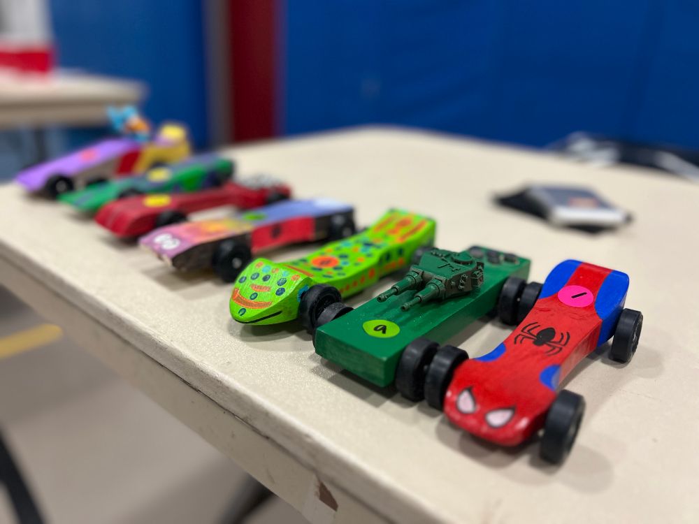 Line of 10 pinewood derby cars. In the foreground is a Spider-Man themed car, then a green tank car, a bright green with polka dots car, a rainbow car. The rest are blurred in the background.