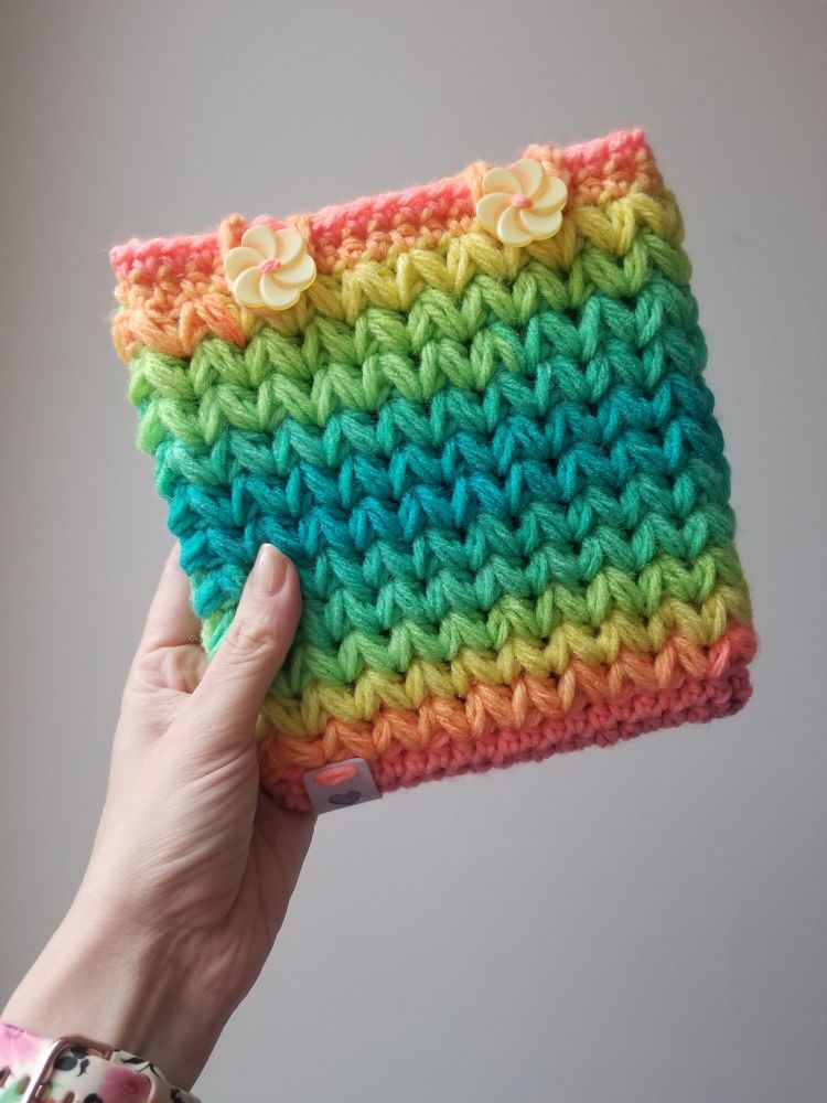 A hand holds a crocheted ereader sleeve in a gradient pastel rainbow with two yellow flower buttons holding it closed