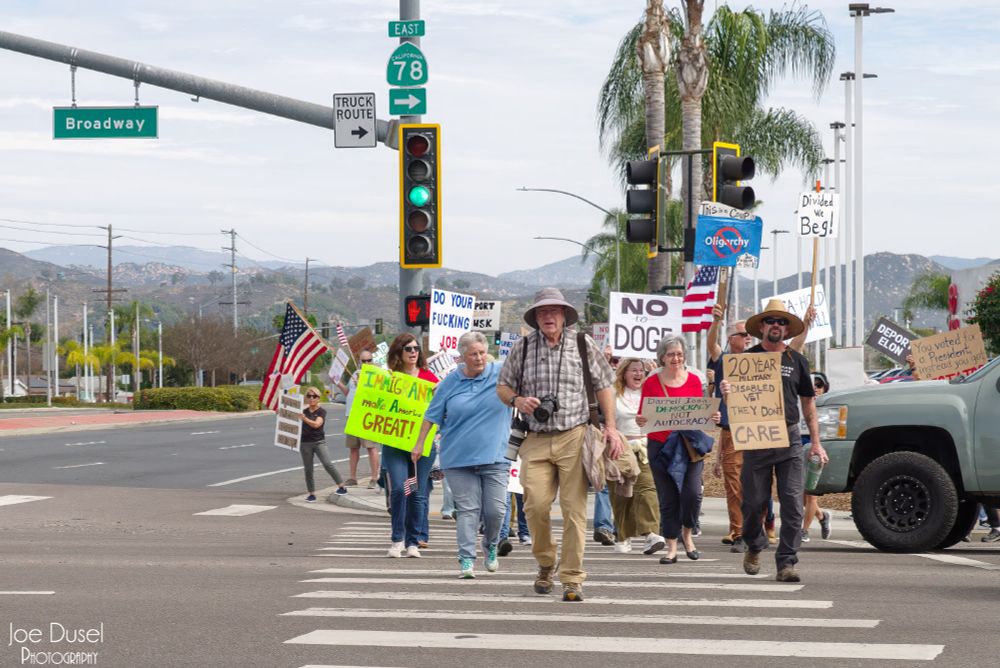 A press photographer with the San Diego Union Tribune accidentally leads a group of protesters crossing the street at a protest against Congressman Darrell Issa in Escondido, CA on Feb 28, 2025. Joe Dusel Photography. 