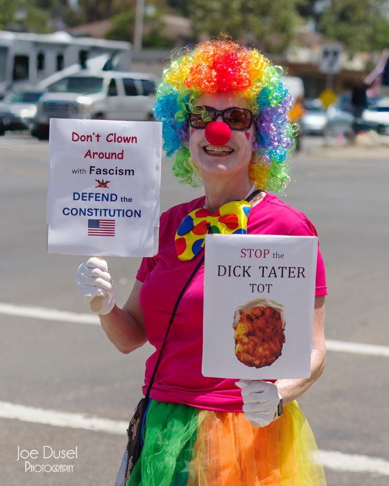 A woman in a clown outfit waved two signs to show her displeasure with the Trump regime at a No Kings protest in Vista, CA on June 14, 2025. Joe Dusel Photography. 
