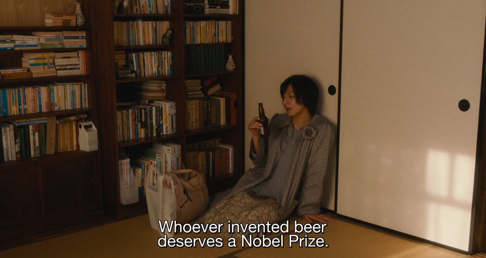 a screenshot from the movie 'Close-Knit' in which Rinko, one of the characters, is drinking beer while sitting on the floor of her apartment. Next to her we can see a bookcase full of books and two groceries bag also on the floor. She is looking at the beer bottle after taking a sip and says 