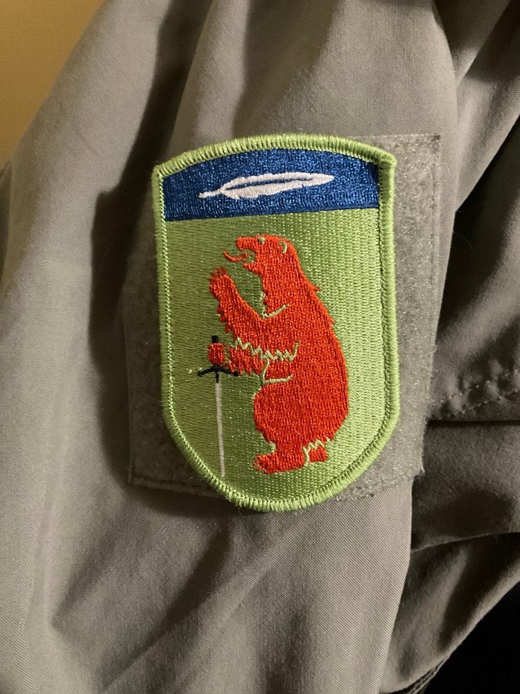 Patch of coat of arms with brown bear holding a claymore sword. Above the bear and sword is a blue field with a white feather. 